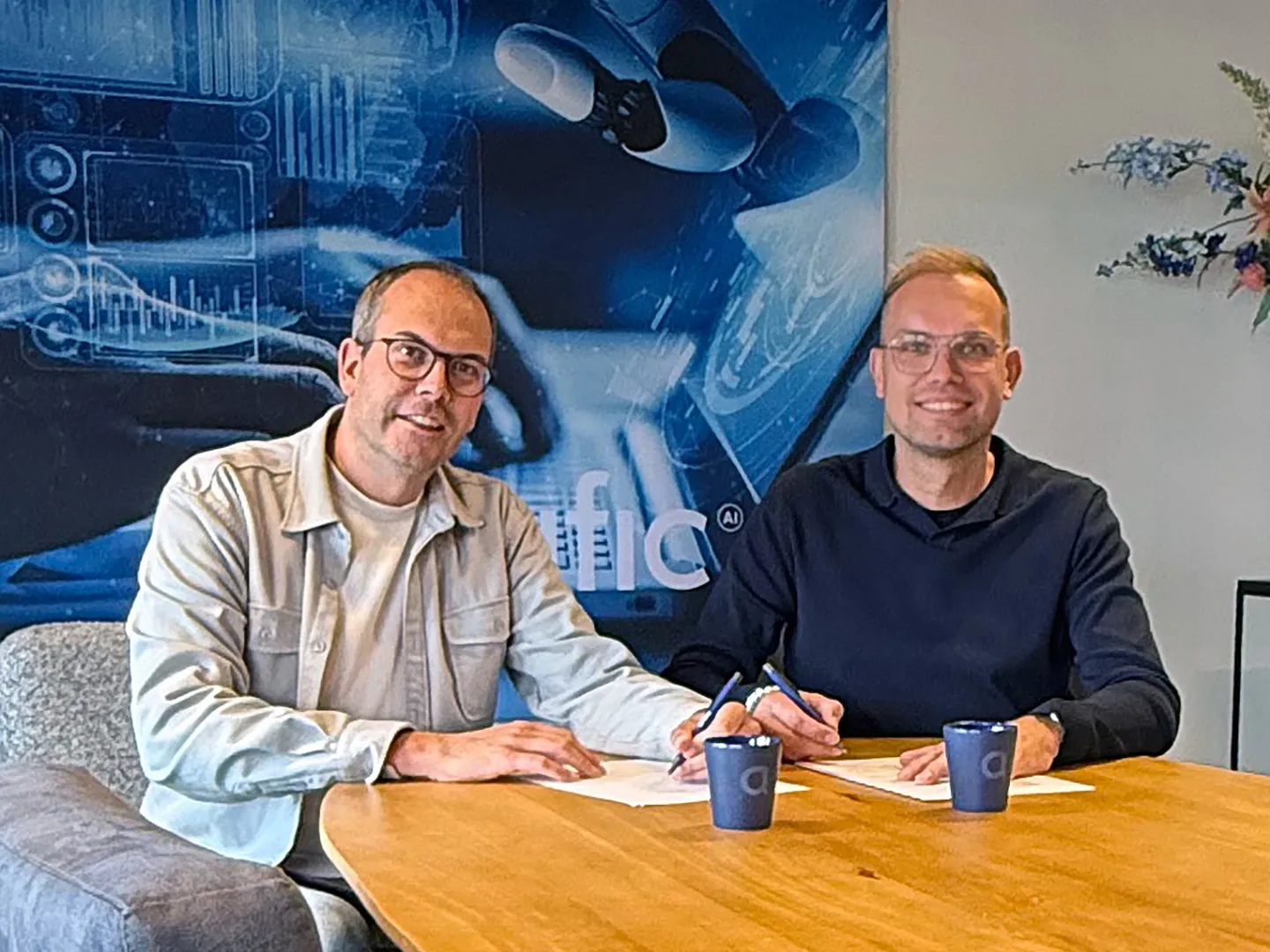 Two men signing papers at a wooden table, in front of a wall poster with the Artific logo.