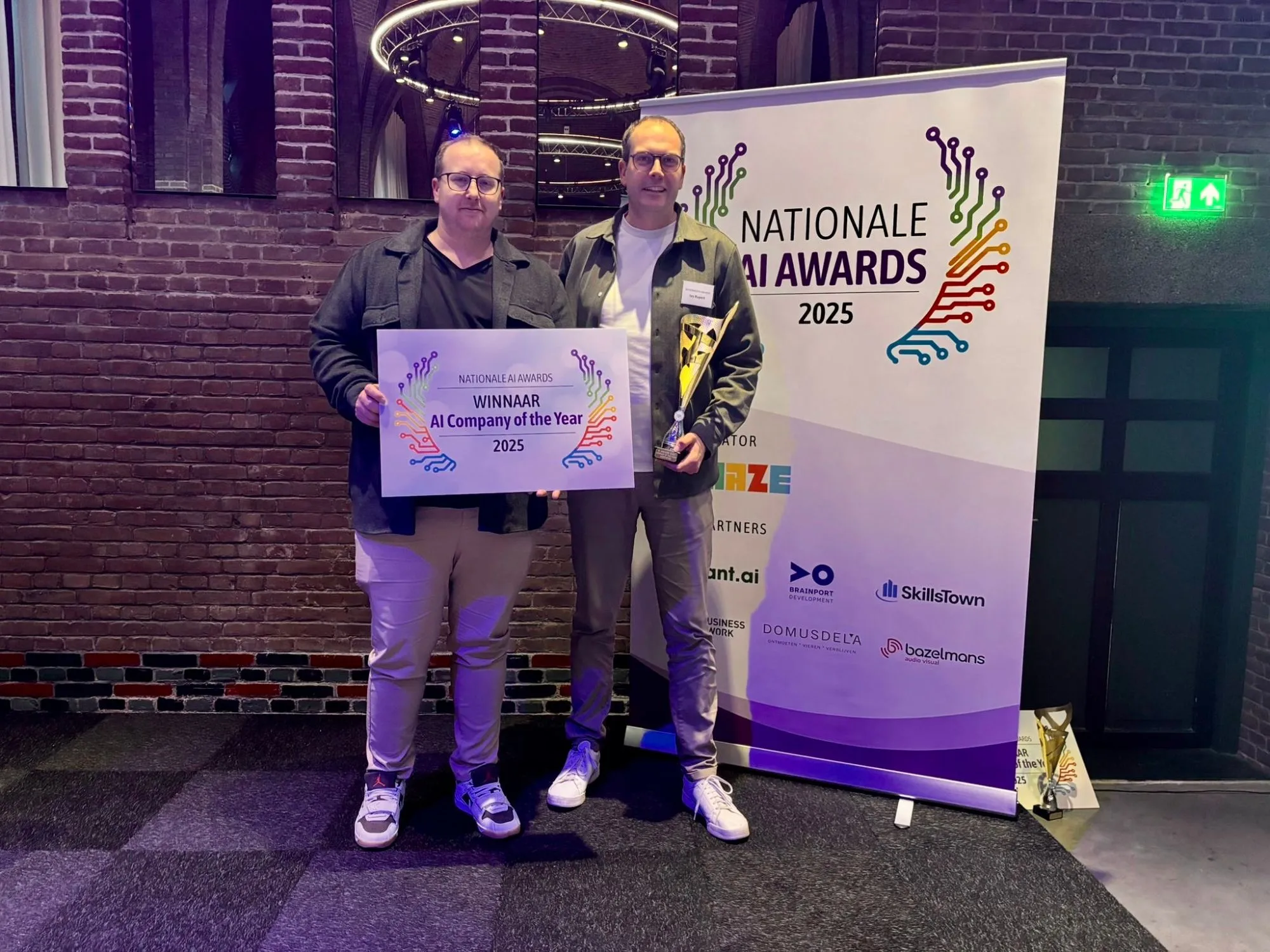 Two men holding a statuette and a sign reading Winnaar AI Company of the Year 2025, in front of a Nationale AI Awards banner.