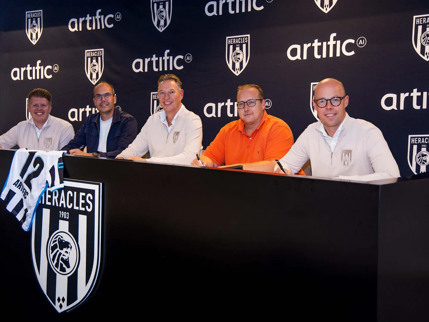 Five men at a table with the Heracles Almelo club crest and a shirt with the number 12, in front of a wall with Heracles and Artific logos.