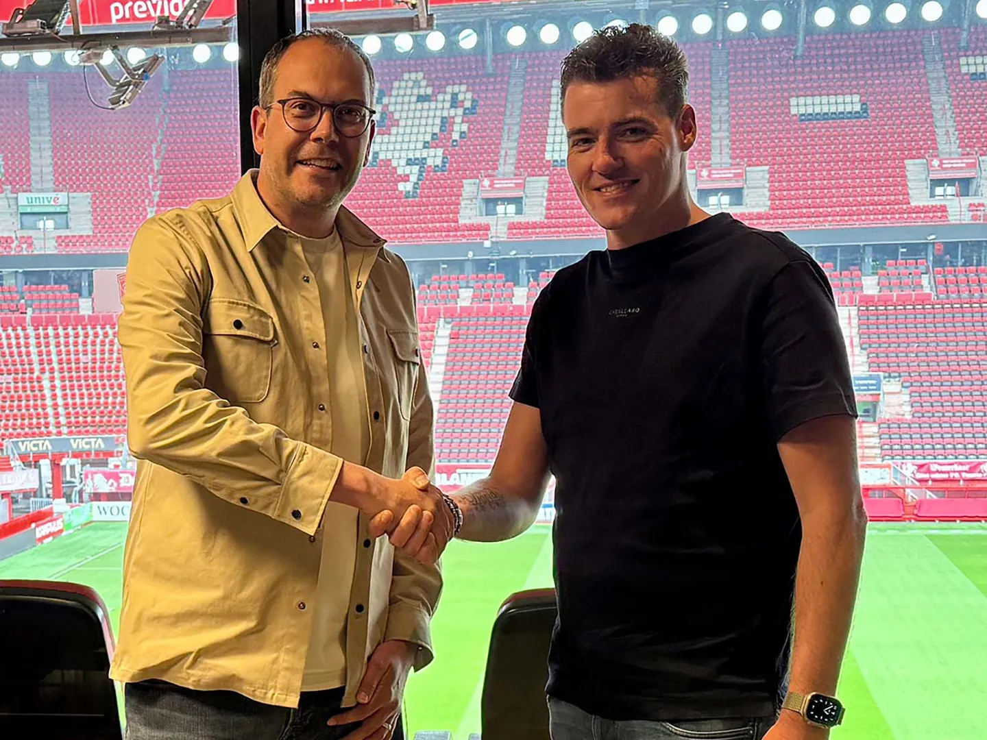 Two men shaking hands in a football stadium, with red stands and the pitch behind them.
