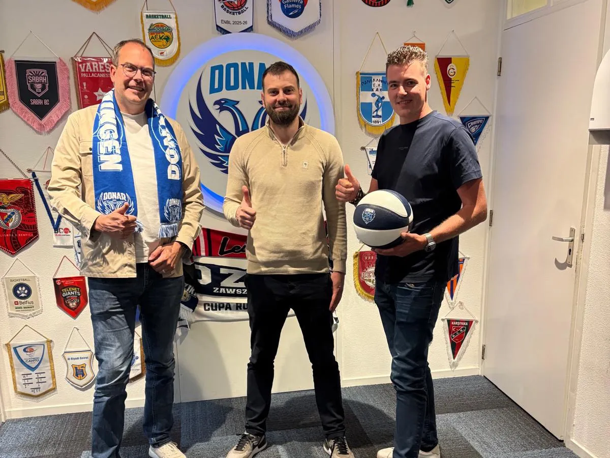 Ivo Rupert mit einem Donar-Schal, Stefan Mladenovic und Erik Davids mit einem Basketball zeigen den Daumen nach oben vor dem beleuchteten Donar-Logo, umgeben von Wimpeln europäischer Basketballclubs.