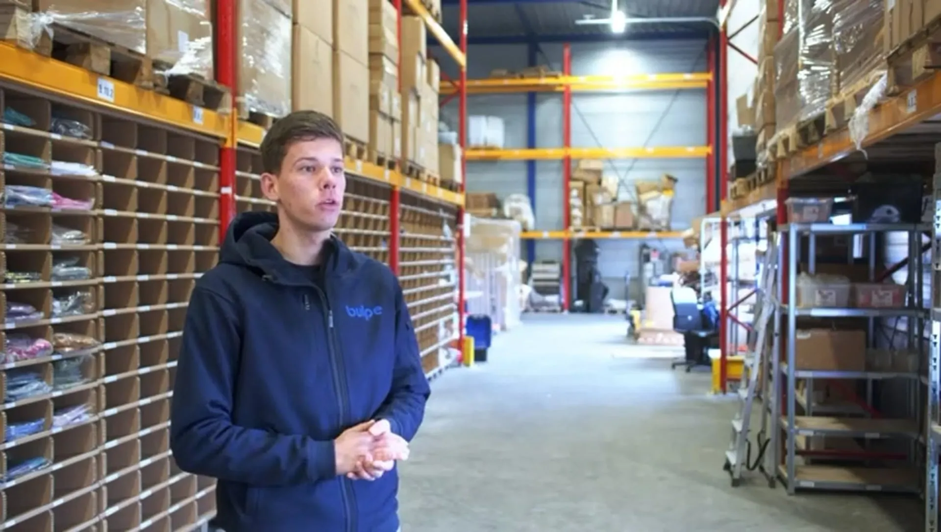 Co-owner Thijs Bult stands between the racks in the Bulpe Fulfilment warehouse and talks, with boxes and order bins behind him.