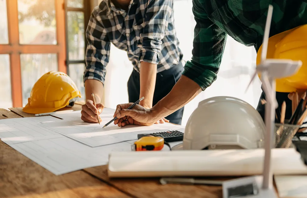 Two people lean over a construction drawing, with hard hats and a model of a wind turbine on the table.