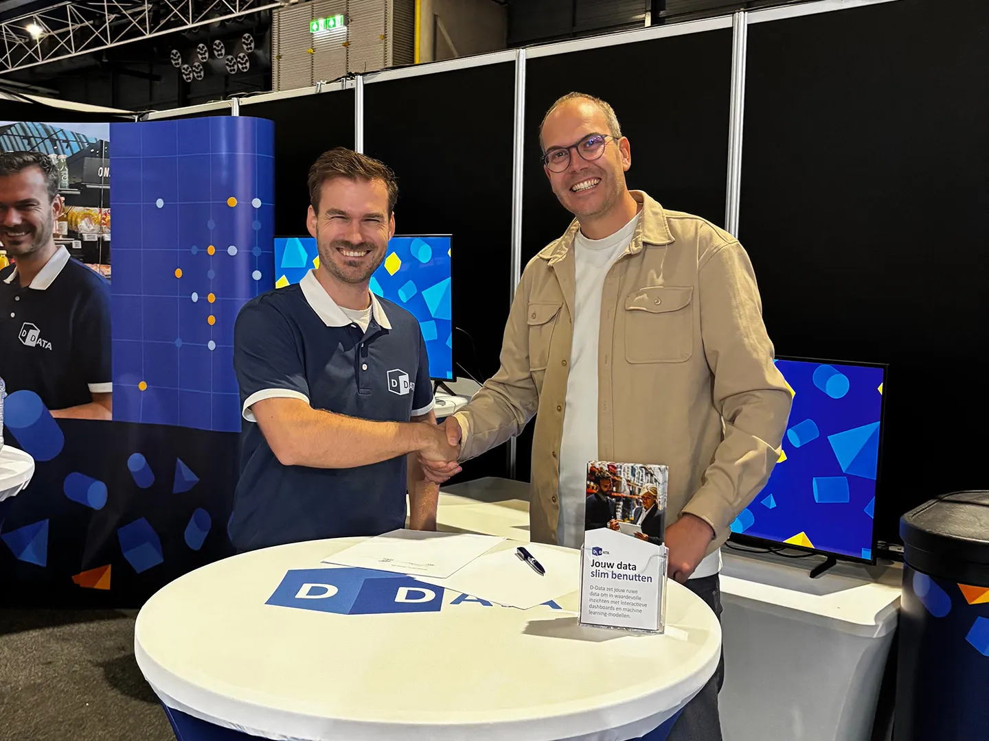 Two men shaking hands at a high table on a D-Data exhibition stand.