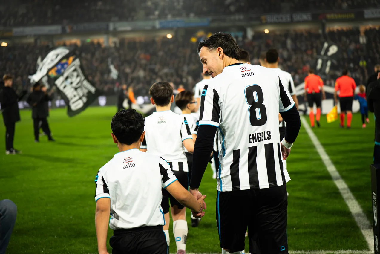 A Heracles Almelo player walks onto the pitch hand in hand with a young supporter, in front of a packed stand.