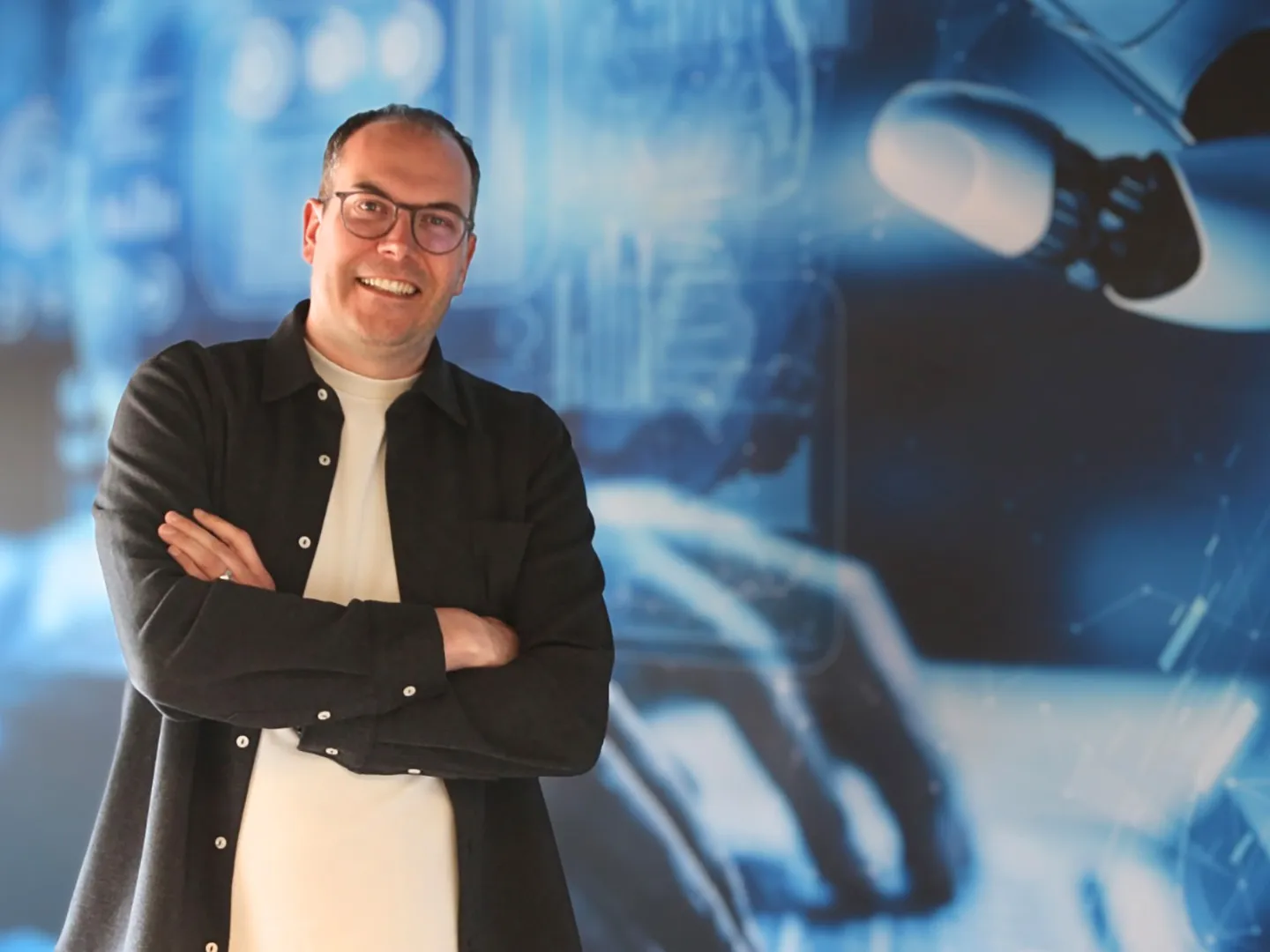 Ivo Rupert with his arms folded in front of a blue wall with a robot hand and a keyboard.