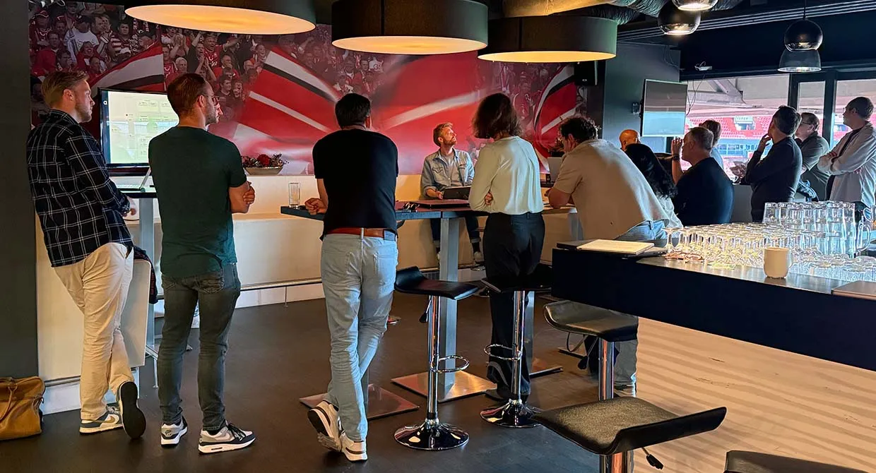 Participants in the partner training stand around high tables in a skybox at De Grolsch Veste, looking out over the stadium.