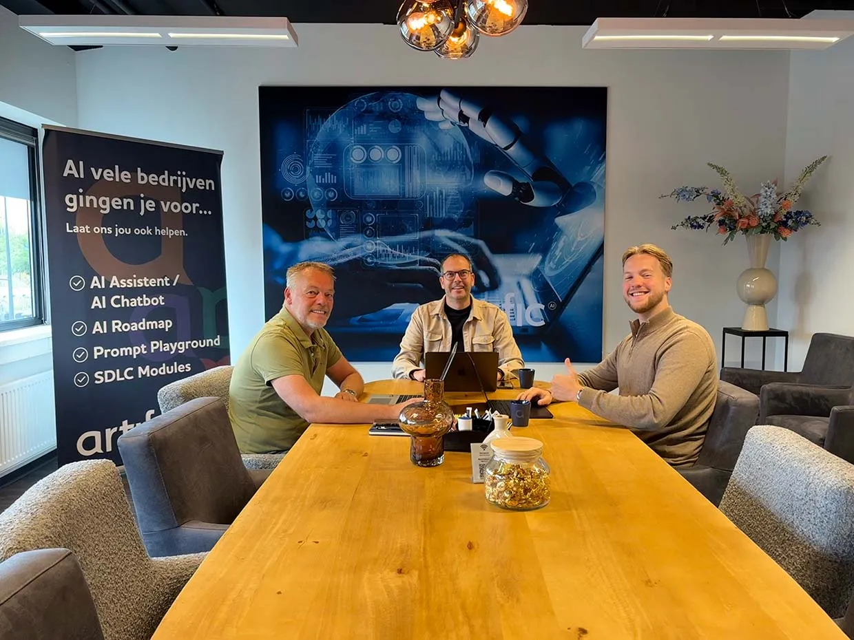 Three colleagues at a long wooden meeting table at Artific, with a banner and a large blue artwork on the wall behind them.