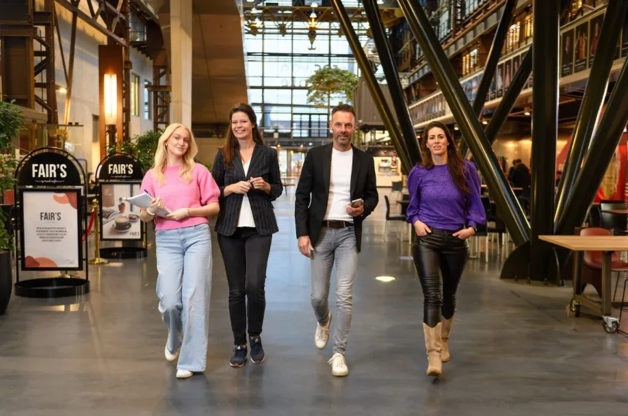 Four ROC van Twente employees walk together through a tall, bright hall with steel trusses.